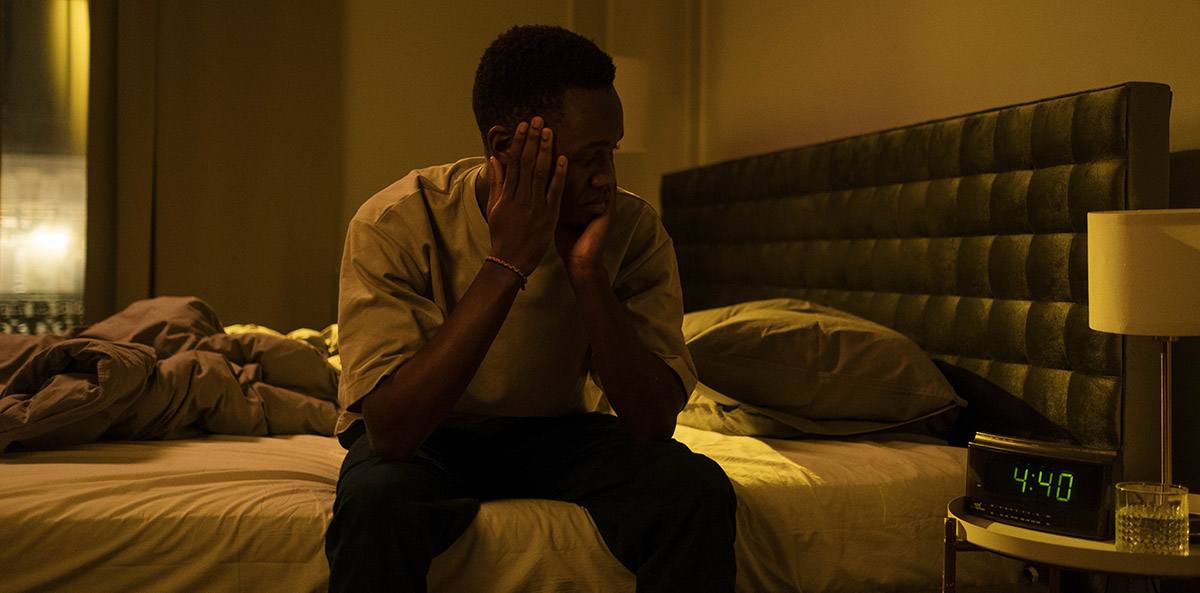 Man sitting on the edge of his bed with his head in his hands, staring at an alarm clock late at night because he's unable to sleep due to Daylight Saving Time