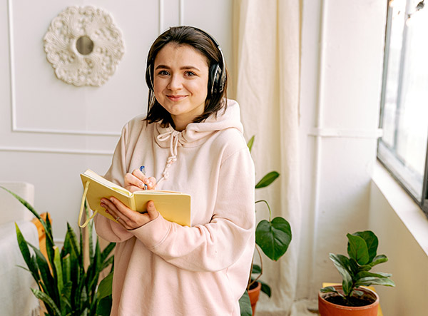 Woman in a pink hoodie listening to music and journaling to relax, de-stress, and boost her mood