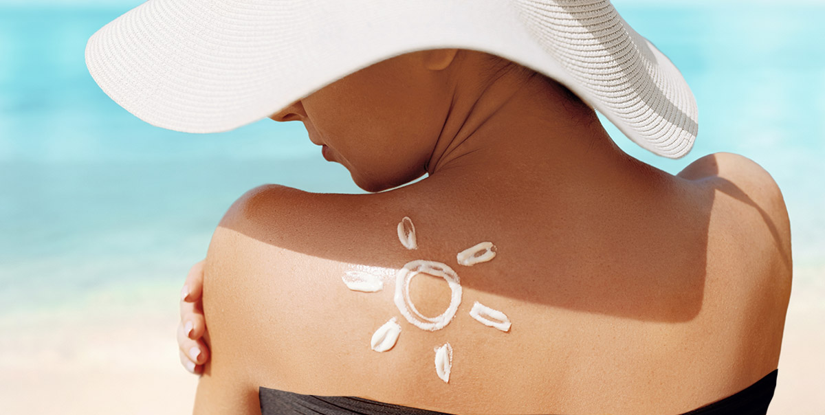 Woman standing on beach in large sunhat with a sun-shaped design of sunscreen on her shoulder
