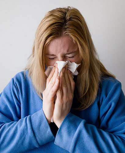 Sick blond woman in blue sweatshirt blowing her nose into a tissue