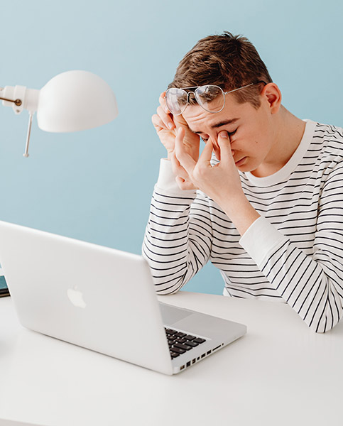 Tired man in a striped shirt pushing up his glasses and rubbing at his eyes because he's feeling rundown while working on his laptop after the Daylight Saving Time clock change