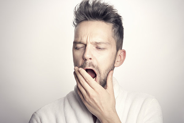 Tired man with bedhead yawning widely and covering his mouth with his hand as he struggles to adjust to Daylight Saving Time