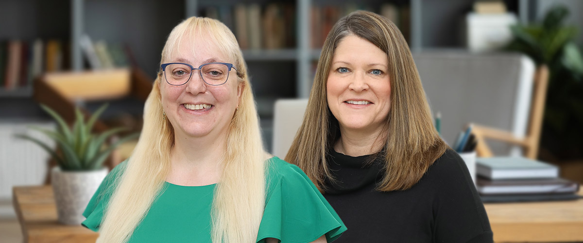 Laurel Health employees Lori Covell, HR Manager, and Susan Usavage-Rudy, Recruiter, standing in front of a desk in an office featuring a bookcase, laptop, and potted plant.