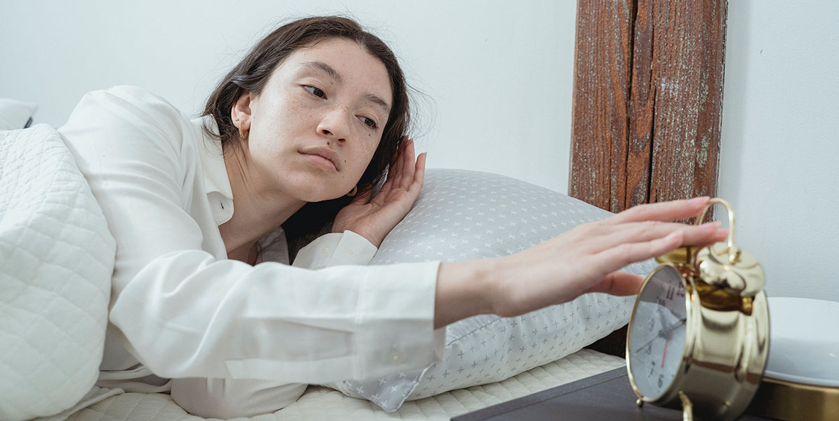 Woman reaching for her alarm clock feeling groggy because her sleep routine has been disrupted by Daylight Saving Time; photo by Miriam Alonso on Pexels