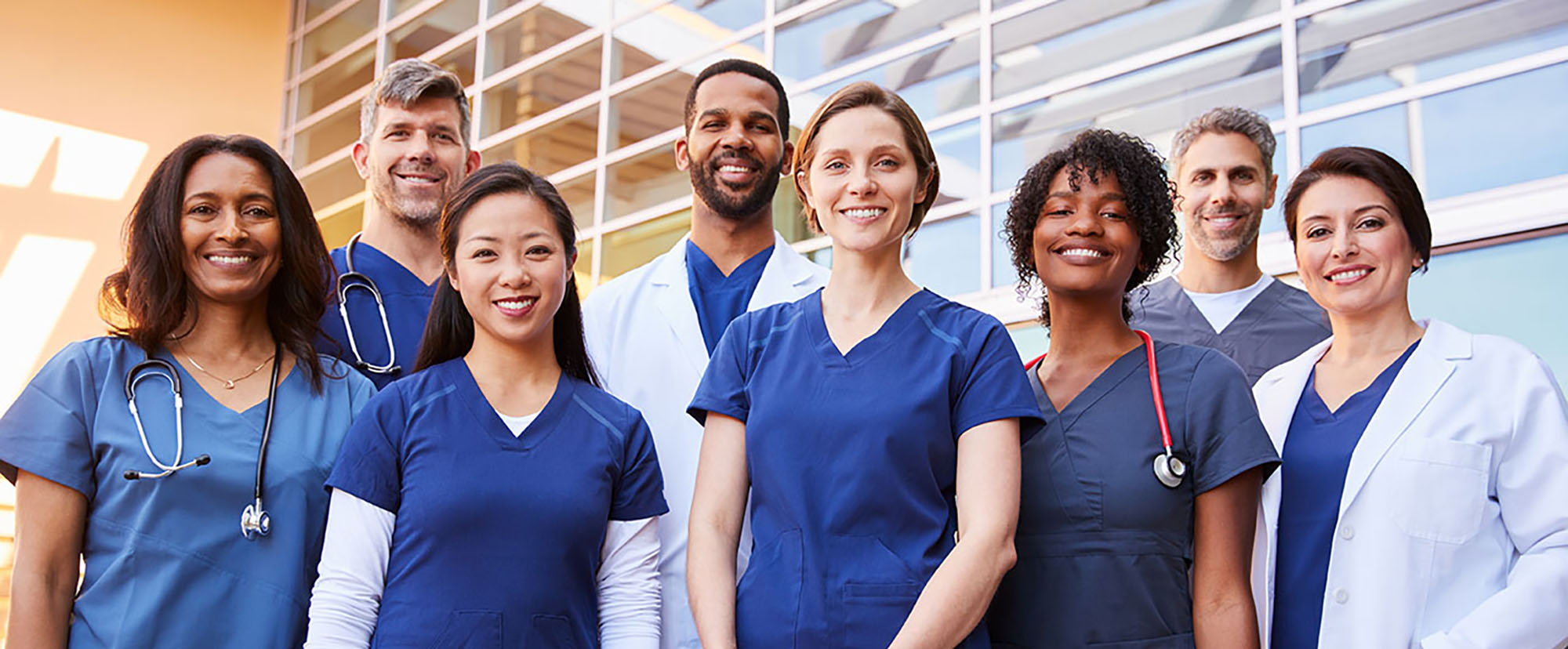 A group of medical professionals standing in front of a glass building smiling and wearing scrubs. Two are wearing white lab coats and three have stethoscopes.