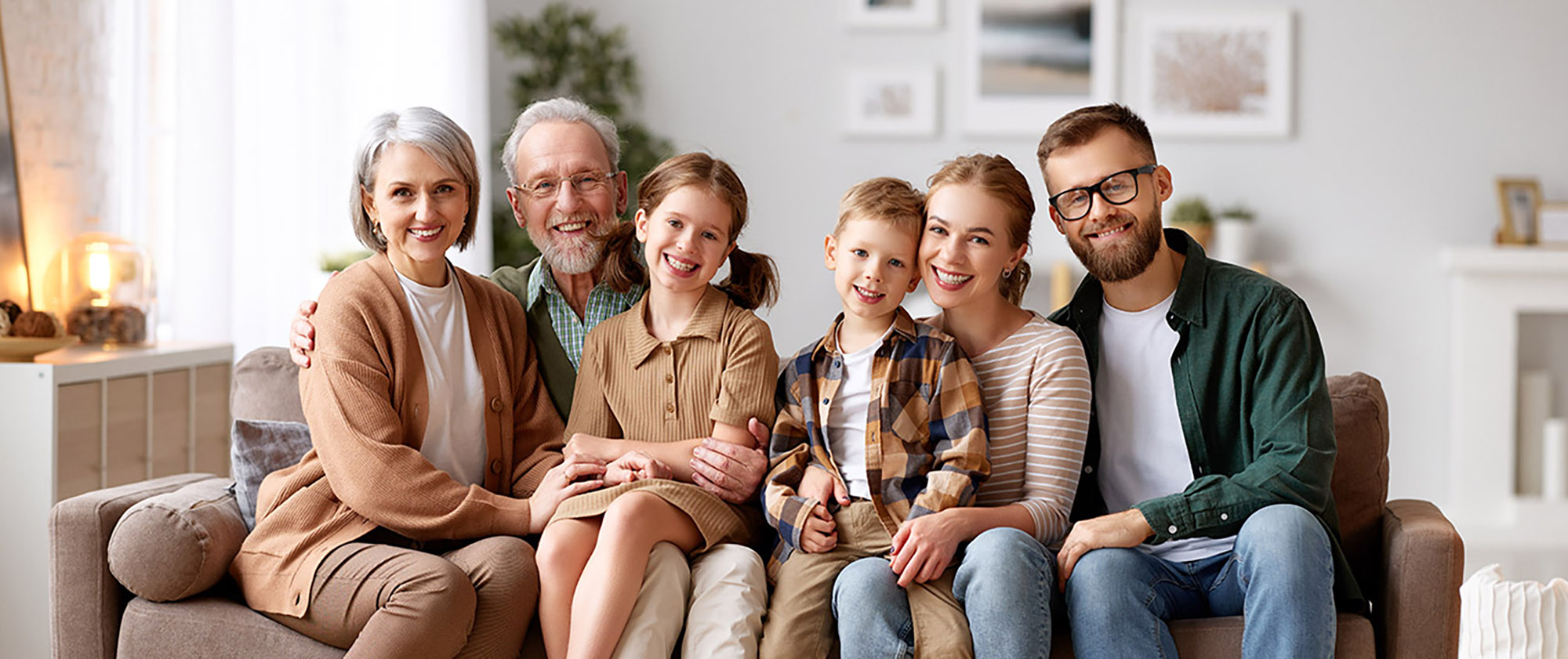 A family sits on a couch smiling at the camera in an inviting living room. The family features two young children sitting between their parents and grandparents.
